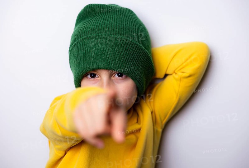 A young child in a bright yellow sweatshirt and green knit hat points forward, showcasing curiosity and playfulness. The backdrop is a plain white wall, highlighting the colors