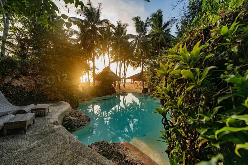 Serene tropical resort pool surrounded by lush green foliage, featuring sun loungers and a backdrop of palm trees and a gazebo, capturing the warm glow of a sunset in the Philippines