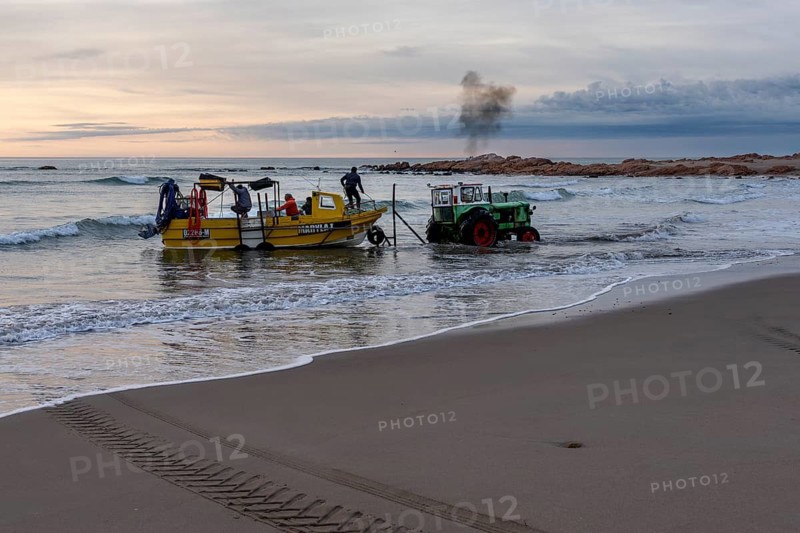Yellow fishing boat with crew is pulled from sandy beach into water by green tractor, rocks in the background, Playa Union, Rada Tilly, Chubut Province, Argentina