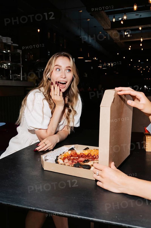 Woman excitedly opening a pizza box at a table, with dim restaurant ambiance. Perfect for visuals related to dining out, pizza delight, or happy experiences