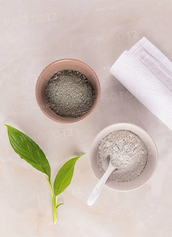 High-angle view of cosmetic gray clay in bowls, adjacent to fresh green leaves and a white towel, ideal for skin care routines
