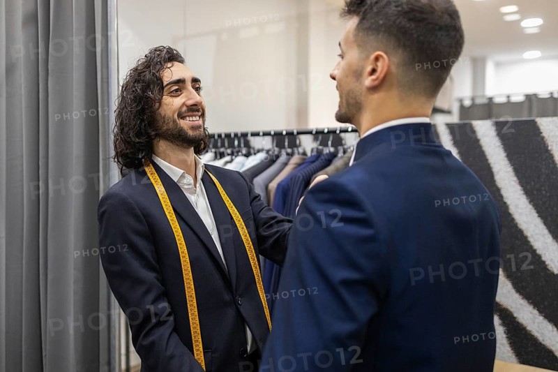 A tailor, measuring tape around his neck, interacts with a client during a suit fitting in a suit store, showcasing professionalism and personal service in menswear fashion