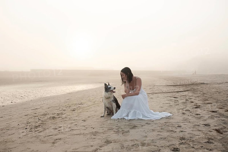 A woman in a flowing white dress sits on a sandy beach with her dog during a tranquil sunrise The misty atmosphere creates an idyllic, peaceful scene