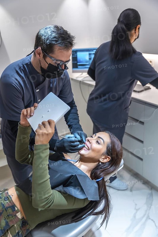 Dentist showing results on tablet to a smiling patient while performing a dental procedure, with a dental assistant working in the background