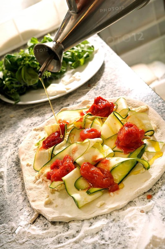 Cropped unrecognizable person drizzling olive oil on a gourmet pizza, topped with zucchini, tomato sauce, and cheese, set on a marble counter