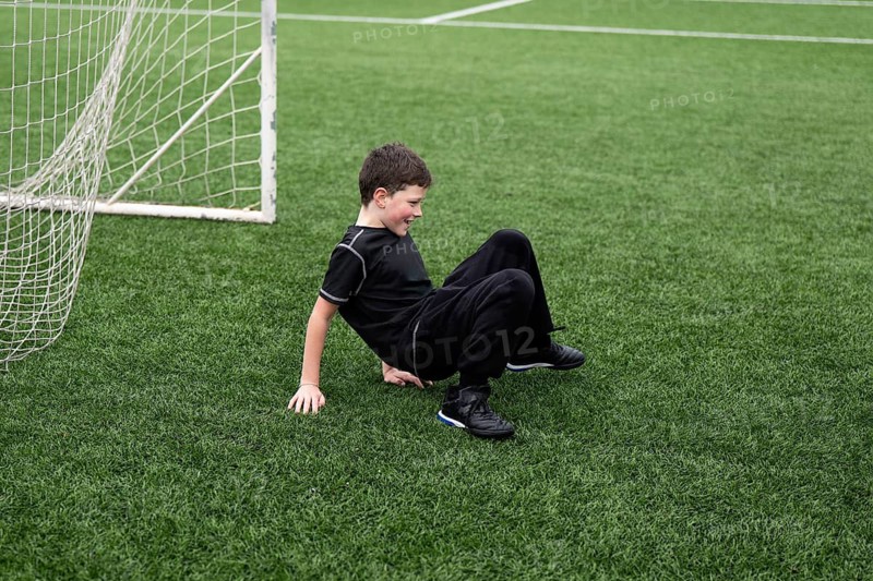A boy dressed in black sportswear smiles on green artificial turf next to a soccer goal, capturing a moment of playful joy during a sports activity