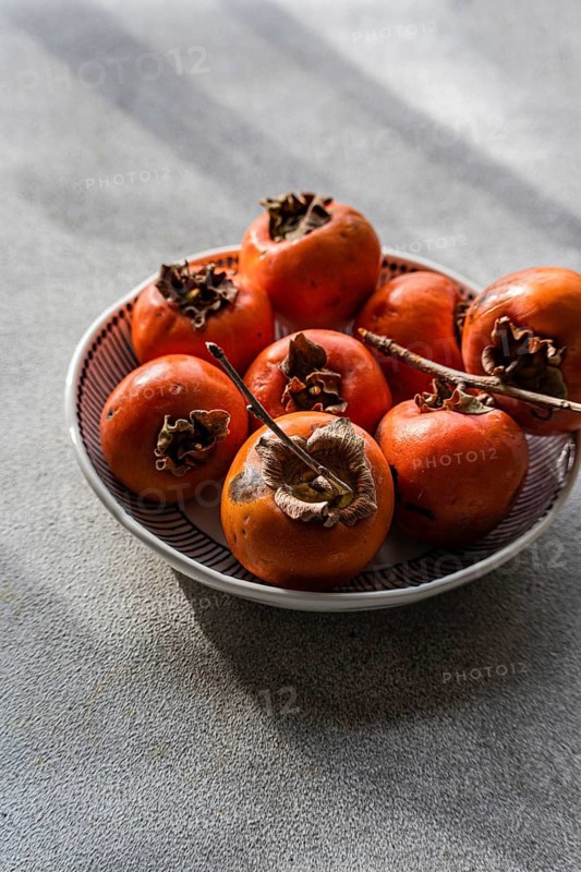 Bowl of fresh, ripe persimmons freshly picked from the orchard, showcased on a subtly textured gray surface, with vibrant natural sunlight enhancing their rich orange color