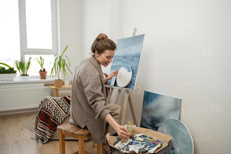 An artist in a cozy, light filled home studio painting a seascape. Surrounded by plants and art supplies, she works with focus and creativity, capturing the sea essence