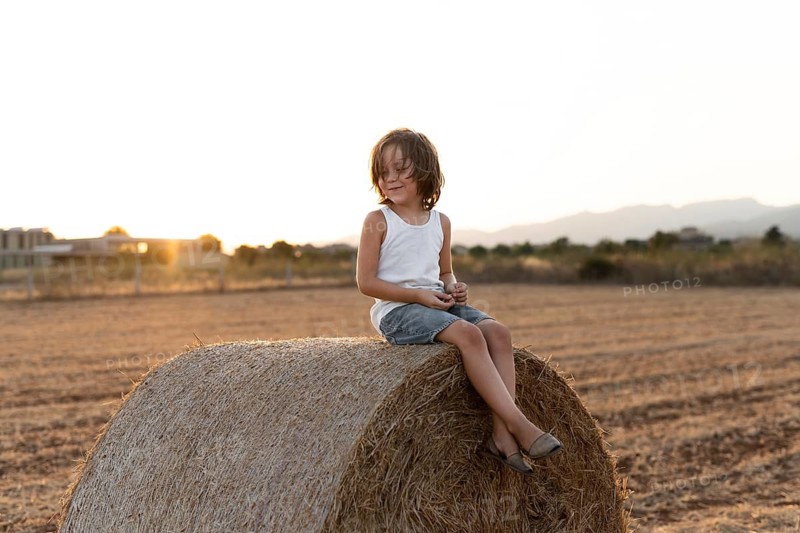 A child in casual attire sits on a large hay bale in a field. The warm sunset in the background enhances the serene rural setting, capturing a moment of youthful freedom and tranquility