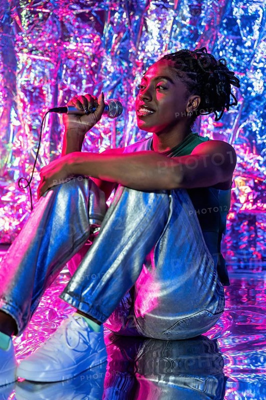 An african woman performer sitting with a microphone, surrounded by vibrant neon colors reflecting off a shiny background, creating an energetic and captivating atmosphere