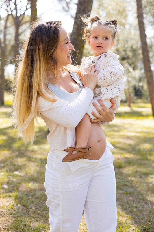 A joyful maternity scene outdoors, featuring a pregnant mother holding her young child. Sunlight filters through trees, highlighting their affectionate bond amid nature