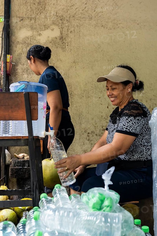 Two asian women prepare fresh coconut water in a rustic setting, using bottles and a pitcher. They work diligently, sharing a smile, surrounded by coconuts and plastic bottles