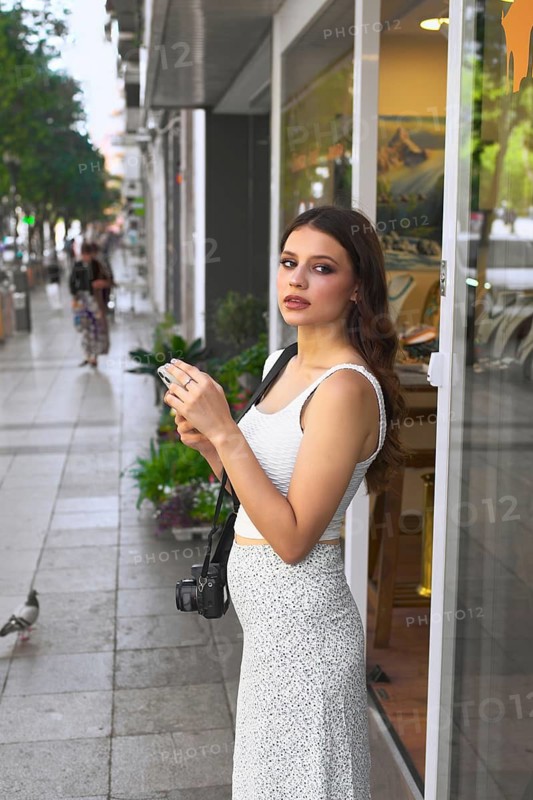 Woman standing outdoors on a city sidewalk holds a phone and wears a camera, ready for photography Trees and shops line the street, creating a lively urban scene