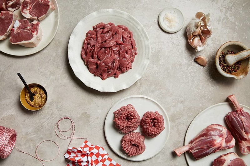 Freshly prepared meat cuts on plates, surrounded by garlic, mustard, colorful peppercorns, salt, and butcher's twine on a rustic surface, emphasizing a gourmet butcher theme