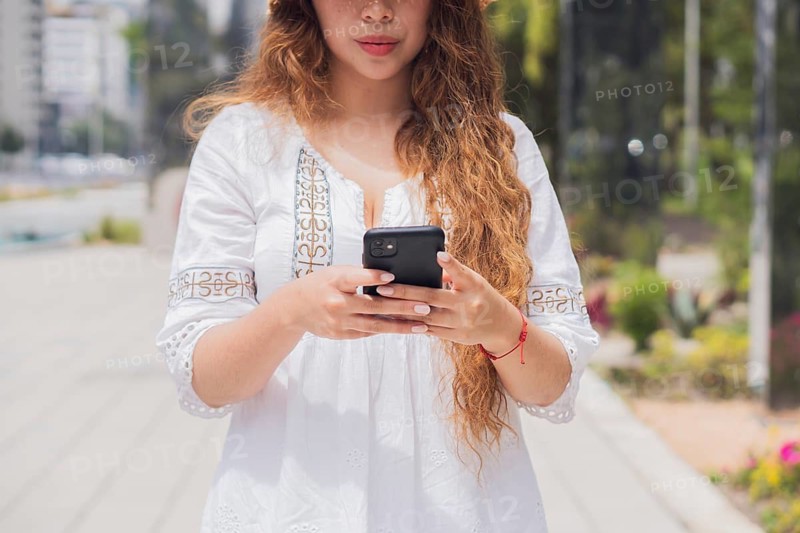 Young American Latin woman with long hair using her smartphone on a sunny day. She wears a white blouse and a bracelet, embodying a modern lifestyle