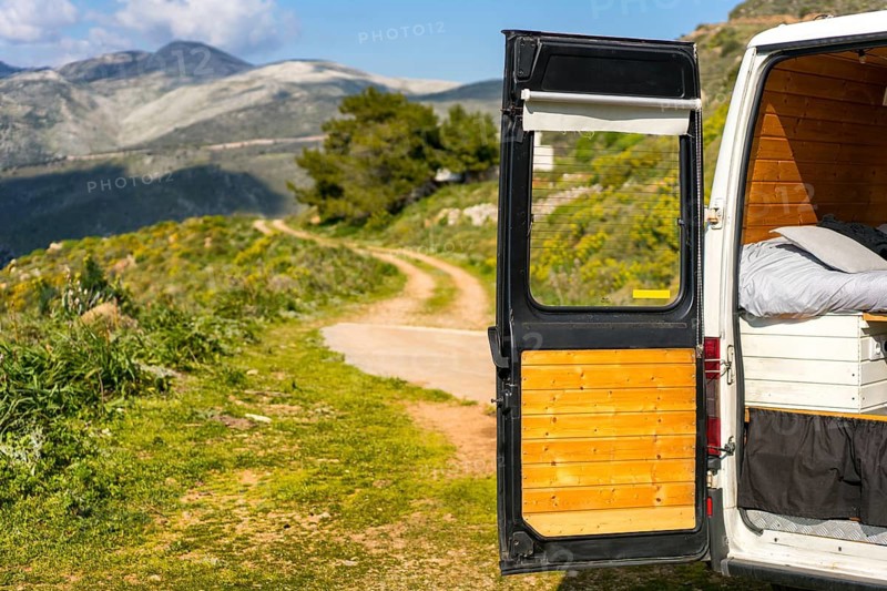 Open door of a camper van reveals a scenic Greek landscape with winding paths and lush greenery, capturing the essence of adventure in a serene setting