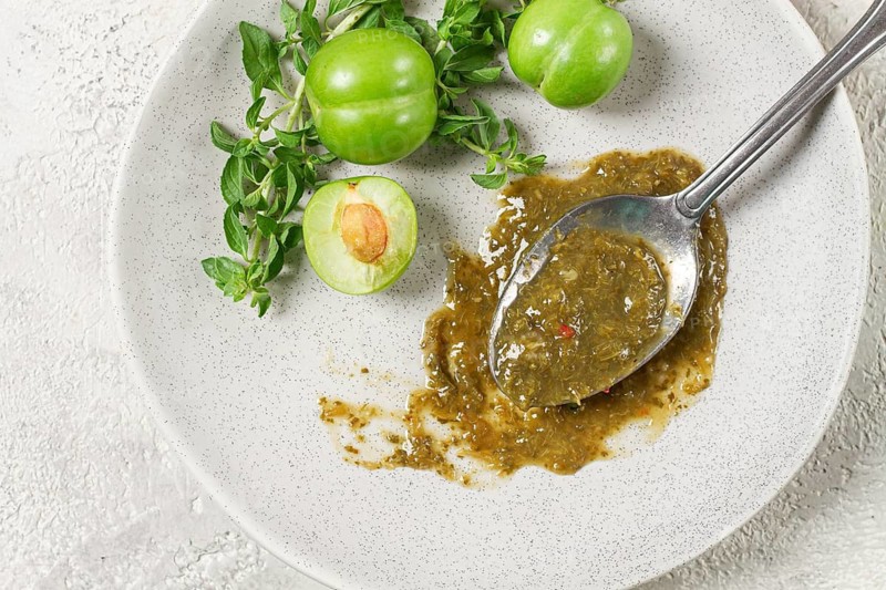 A spoon with tkemali sauce, on a plate, with cherry plum and mint, top view, no people