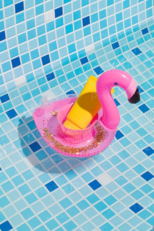 A vibrant yellow sunscreen bottle rests in a playful pink flamingo float on a pool's blue and white tile surface, capturing the essence of summer relaxation