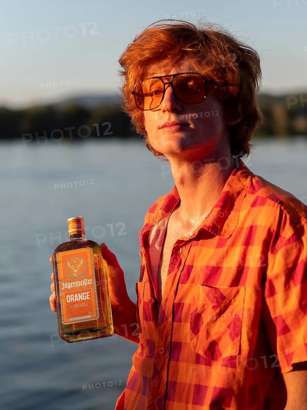 Young man standing by the lake, holding a bottle of alcohol in his hands, red hair, orange shirt, herbal liqueur, portrait