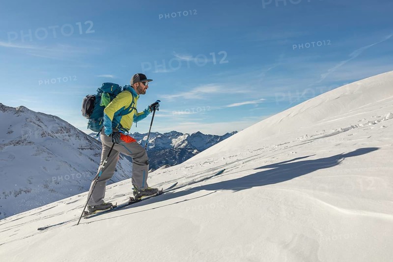 A man ski touring uphill on a pristine snowy mountain, equipped with poles and a backpack under a clear blue sky. Captures adventure and the beauty of winter