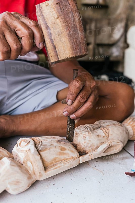 An Indonesian man skillfully carves a wooden sculpture using a chisel and mallet, showcasing traditional artistry and cultural craftsmanship in a detailed close-up shot