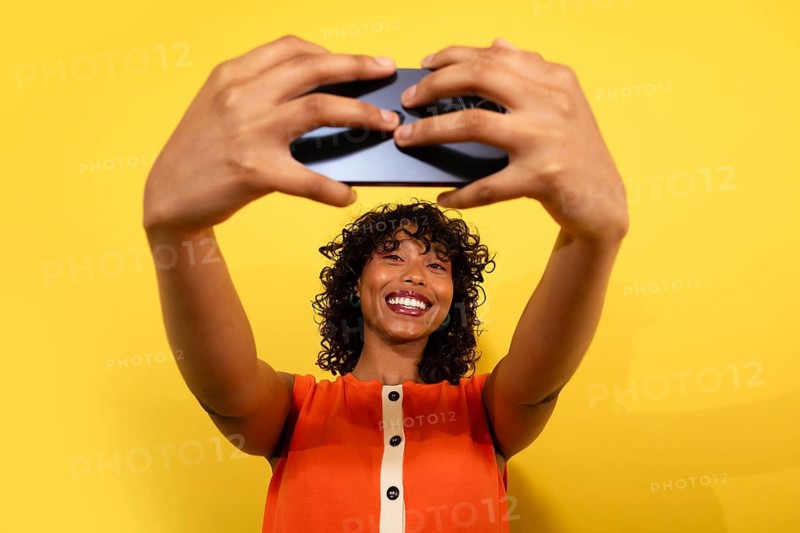 A cheerful woman with curly hair and a bright smile takes a selfie against a vibrant yellow background, capturing a joyful moment with her smartphone in an orange top