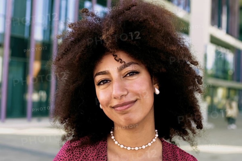 A young Arab woman with a radiant smile and afro hair poses outdoors looking at camera. She wears a stylish pink blouse and pearl jewelry, embodying confidence and beauty in a modern cityscape