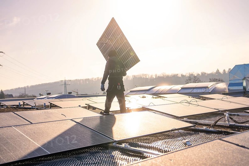 A worker carries a solar panel over a roof at sunset, a hilly landscape in the background, PV installation at OBI Baumarkt, Mühlacker, Enzkreis, Germany