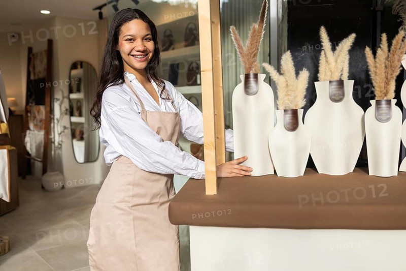 A woman wearing an apron arranges decorative vases on a counter in a chic bag store. Shelves with handbags are visible in the background, creating a cozy retail atmosphere