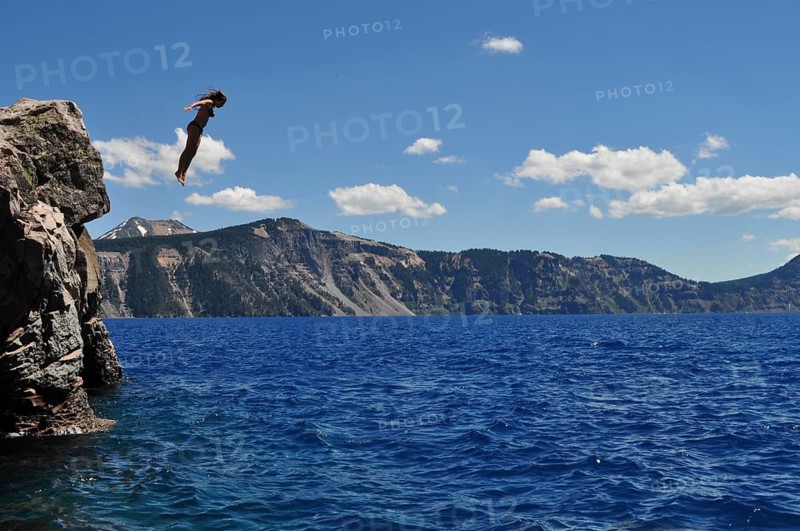 A person jumps from a rocky outcrop into the ice-cold, blue water of Crater Lake, Crater Lake National Park, Oregon, USA
