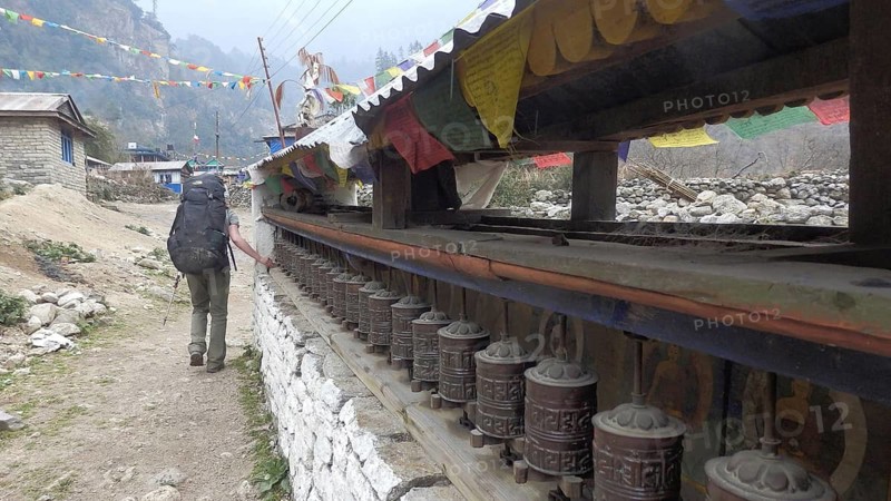 Hiker with large backpack passes Buddhist prayer wheels in a foggy mountain village, trekking at Annapurna Circuit, Manang, Nepal