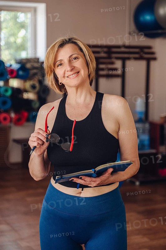 A woman personal trainer stands in a gym, smiling looking at camera while reviewing her planner. She holds a pair of glasses and a pen, reflecting on her fitness schedule and planning client sessions