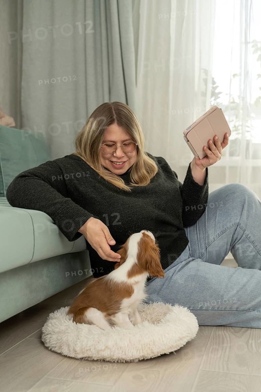 A woman sits comfortably in her living room, smiling at a small dog. She holds a tablet and enjoys a cozy moment with her pet on the floor