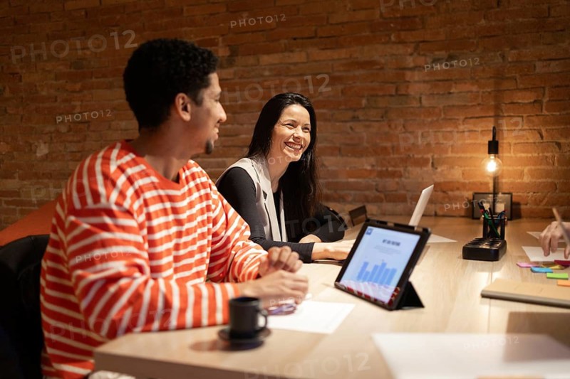 A diverse group of coworkers engage in a collaborative discussion around a modern table. Laptops and tablets show teamwork in a warm, inviting, and creative workspace