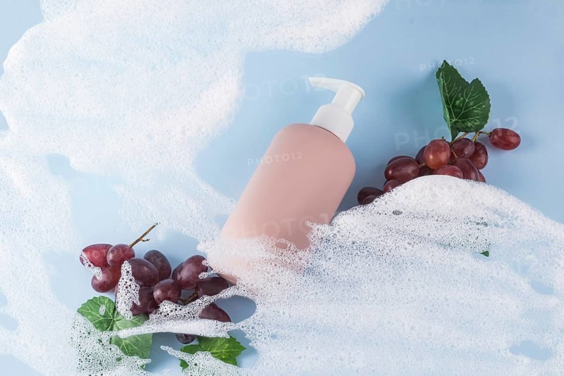 A pink bottle of grape-scented shower gel on a blue background. Surrounded by soap bubbles and red grapes, evoking freshness and indulgence