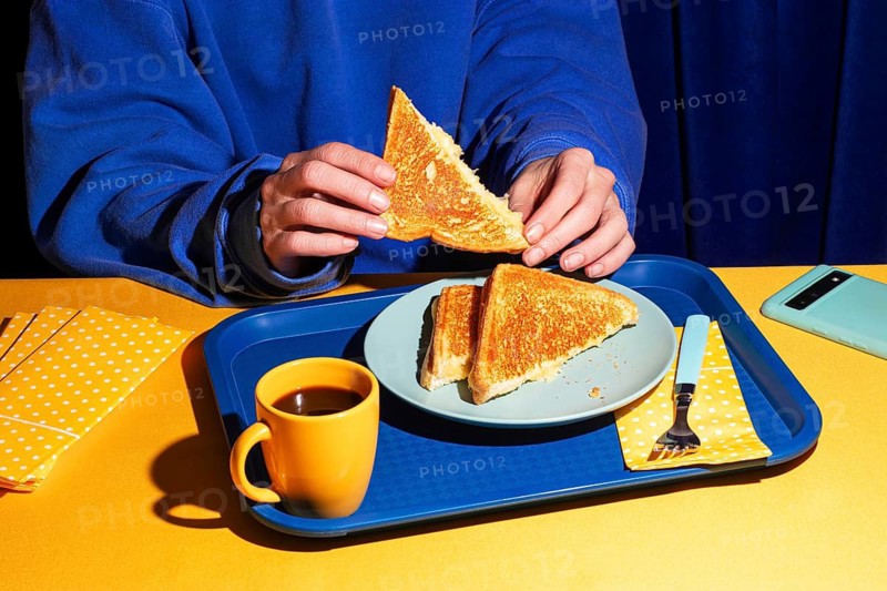 Overhead view of a person in a blue sweatshirt holding a crispy grilled cheese sandwich. Harsh light and vibrant colors enhance the contrast