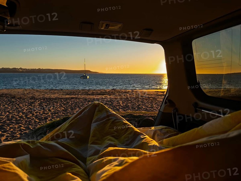 Relax and enjoy a serene sunrise view from the comfort of a camper van by the beach in Phillip Island, Australia. A sailboat drifts on the calm sea, creating a perfect setting for tranquility