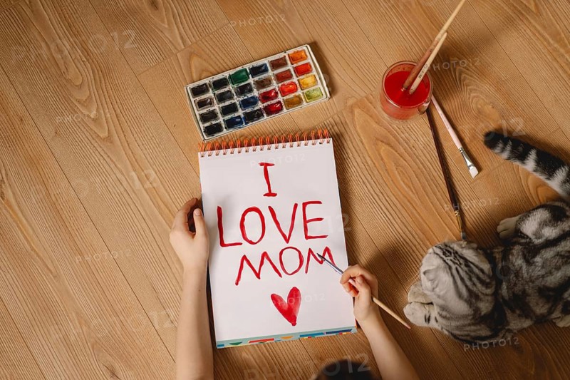 A young boy uses watercolors to create a heartfelt Mother's Day card. With a palette of colors and a loving message, he expresses his affection on paper, accompanied by a curious cat