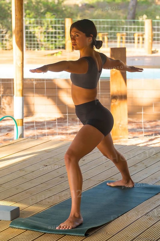 Young woman practicing virabhadrasana ii yoga pose on a mat outdoors, showcasing balance and flexibility amid a tranquil natural setting, embracing fitness and wellness