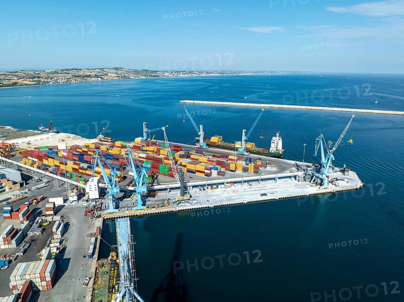 Aerial perspective of Ancona's bustling port, showcasing colorful cargo containers and towering cranes. The vivid blue sea provides a stunning backdrop to this industrial scene