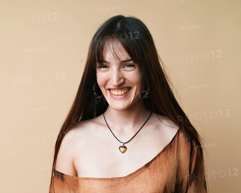 Portrait of a joyful young woman wearing a brown patterned blouse and a necklace, laughing against a beige studio background. Her hair is long and dark, and she displays a natural, pleasant expression