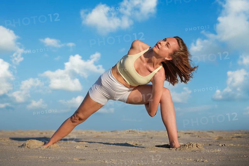 A woman practices Baddha Trikonasana on a sandy beach during sunset with her arms bound behind her back and legs wide apart