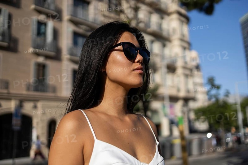 Chinese woman wearing sunglasses and a white dress strolls through a vibrant city during a sunny summer day, reflecting a confident and stylish urban lifestyle