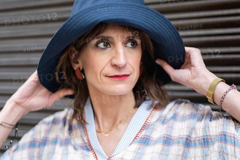 A fashion woman in a plaid outfit, wearing a navy hat, poses confidently against a dark background. Her stylish appearance and calm expression evoke elegance