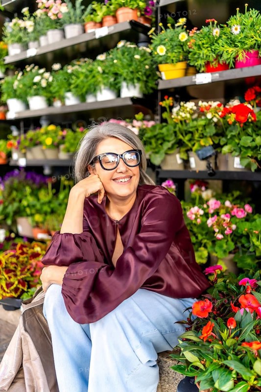 Happy woman with glasses and grey hair sitting in a vibrant flower shop, enjoying the colorful plants and a beautiful smile, representing joy, retail, and natural beauty