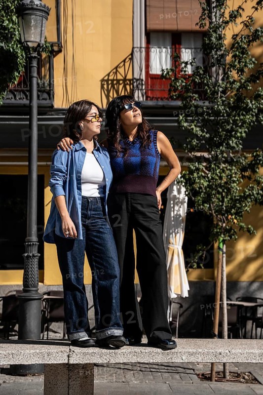 A multiethnic lesbian couple stands arm in arm, enjoying a sunny day in the city. They exude happiness and pride, showcasing love and diversity in an urban setting