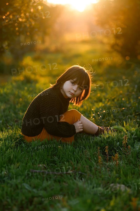 A young child sits on vibrant green grass with orange trees, bathed in the warm, golden glow of a sunset. The peaceful setting and natural light create a serene and nostalgic atmosphere