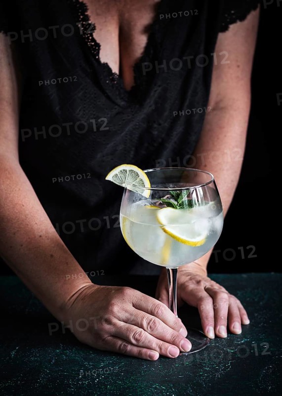 A woman wearing a black dress holds a refreshing gin tonic garnished with lemon slices and mint. The elegant, chilled drink sits on a dark tabletop