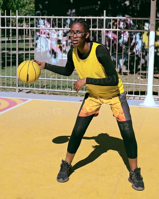 A woman athlete in sporty attire practices basketball on a vibrant outdoor court. Wearing a bright yellow vest, the player focuses on dribbling skills under the sunlit sky