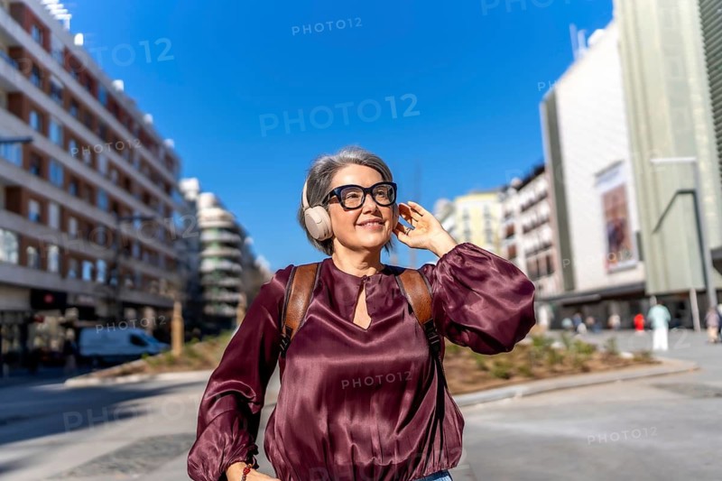 Stylish middle aged woman with gray hair and glasses walks confidently along a sunny city street, smiling and enjoying music through headphones, relaxed and independent